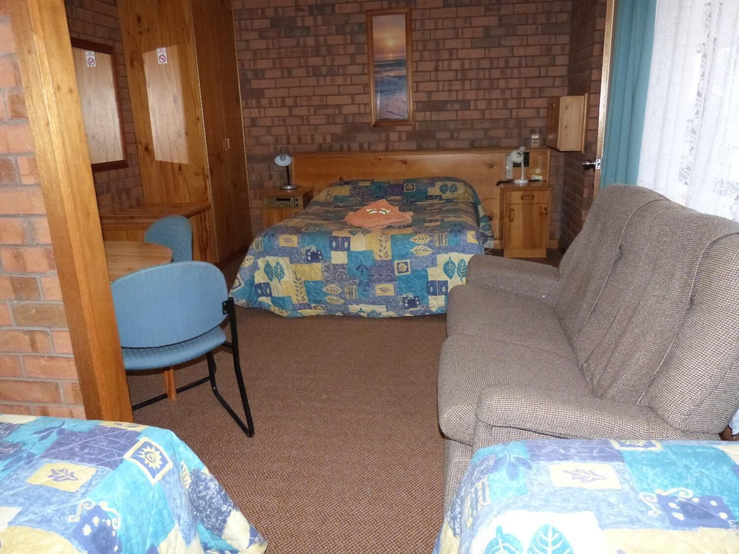 A room with a brick wall, a bed with a colorful quilt, a grey sofa, a blue chair, and a wooden cabinet.