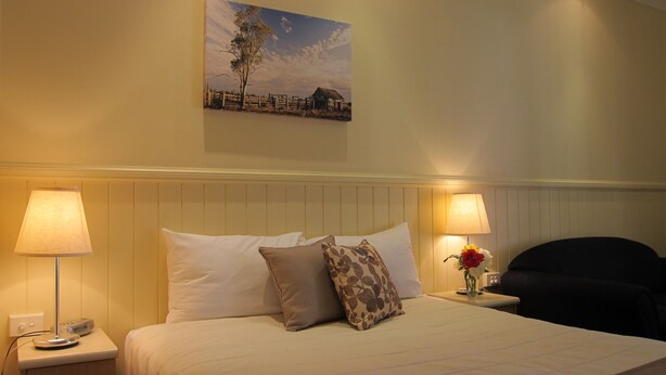 Habitación de hotel con una cama, dos lámparas de noche, una mesita de noche con un teléfono, una silla, un cuadro en la pared y un jarrón con flores.