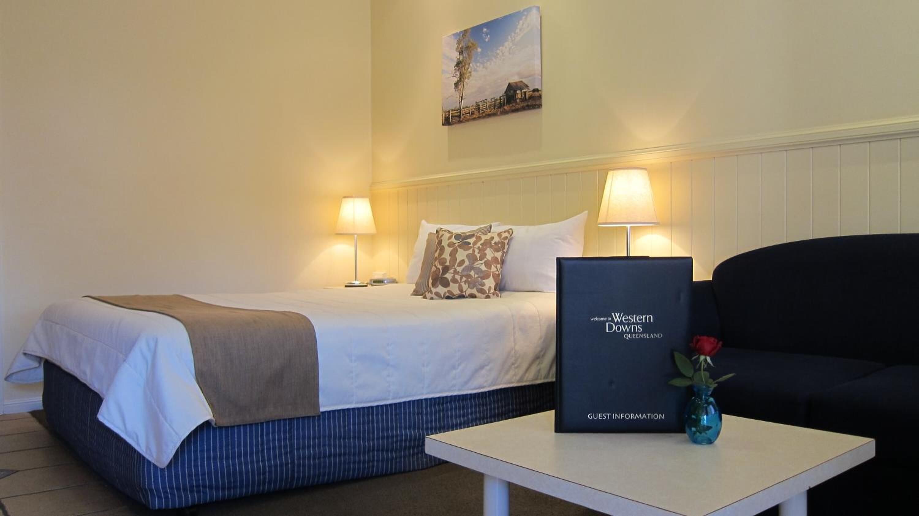 A hotel room with a bed, two bedside lamps, a framed welcome sign, a small table with a vase, and a painting on the wall.