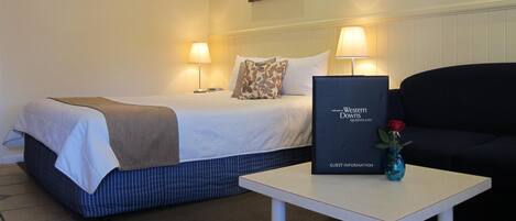 A hotel room with a bed, two bedside lamps, a framed welcome sign, a small table with a vase, and a painting on the wall.