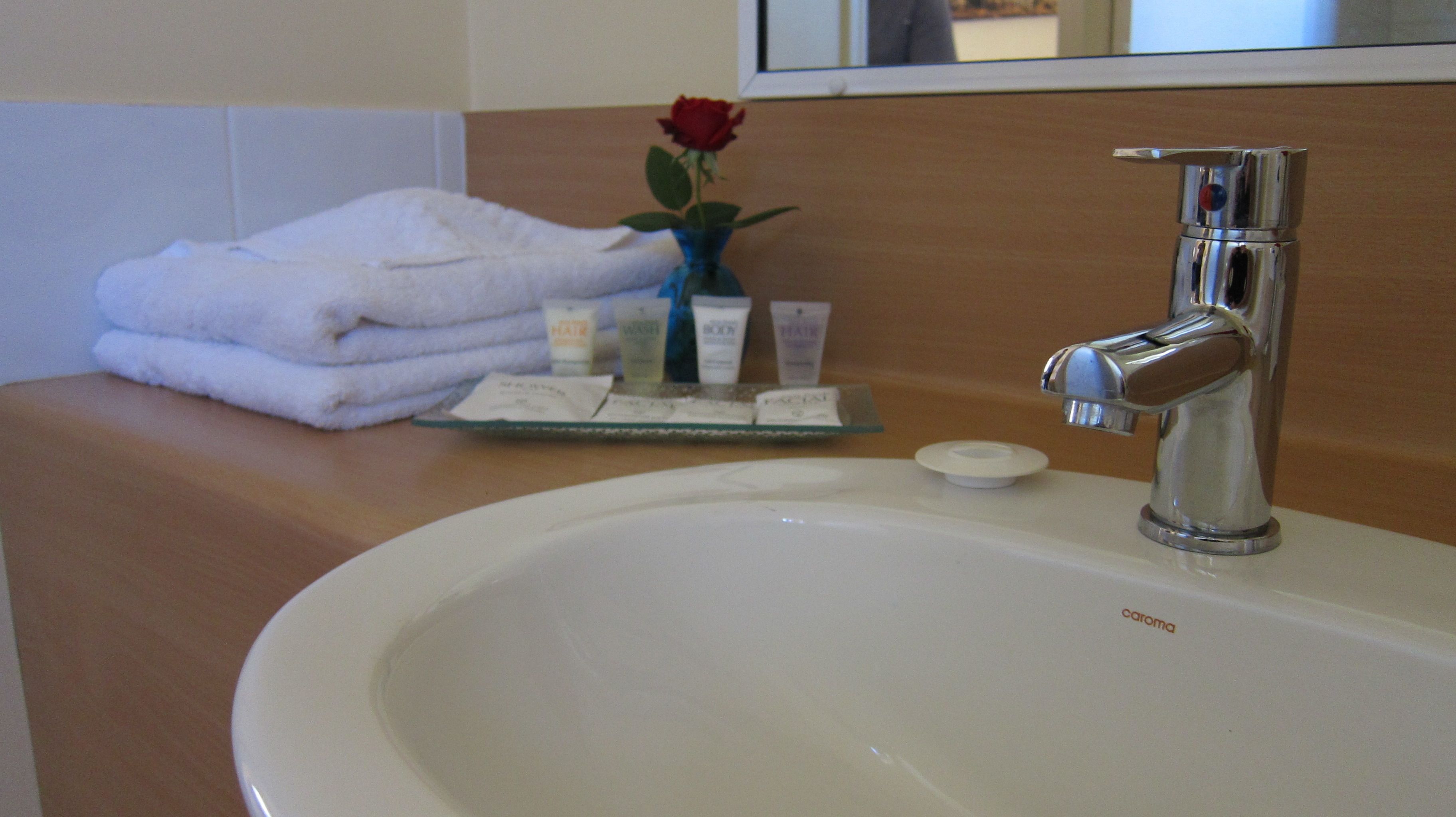 Baño de hotel con lavamanos, artículos de aseo y una rosa en un jarrón.