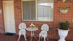 Edificio de ladrillo con puerta blanca, ventana con mosquitera y mesa exterior blanca con dos sillas. Hay una planta en maceta a la derecha.