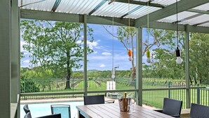 Un patio cubierto con una mesa de madera y sillas de mimbre, con vistas a una piscina y un jardín cercado.