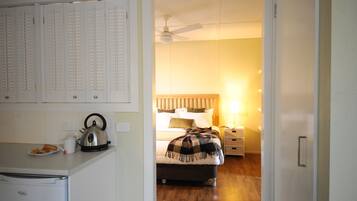 Quarto com cama, mesa de cabeceira e pequeno espaço de cozinha equipado com chaleira e um prato de comida.