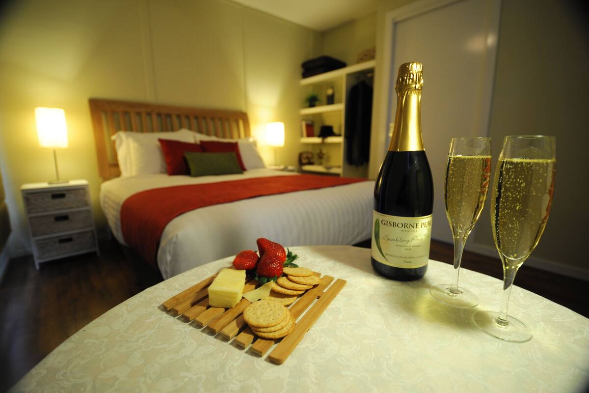 Habitación de hotel con una cama bien hecha, una mesita de noche con lámpara, una botella de champán y copas sobre una mesa.