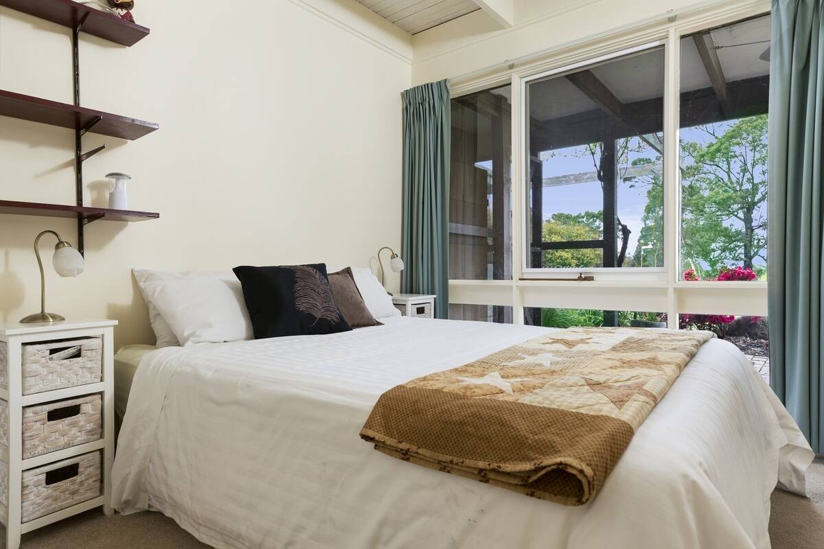 Un dormitorio con cama, mesita de noche, lámpara, ventana con cortinas y vistas a árboles.