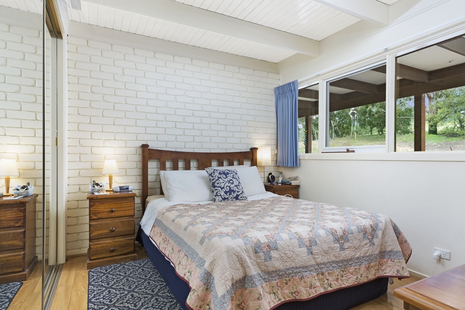 A bedroom with a brick wall, a wooden bed, a nightstand, a bedside table, a window with a view of greenery, and a patterned rug.