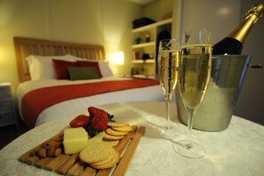Bandeja de desayuno ordenada con fresas, queso y galletas sobre una cama con una colcha roja, junto con dos copas de champán y una botella en un cubo de metal.