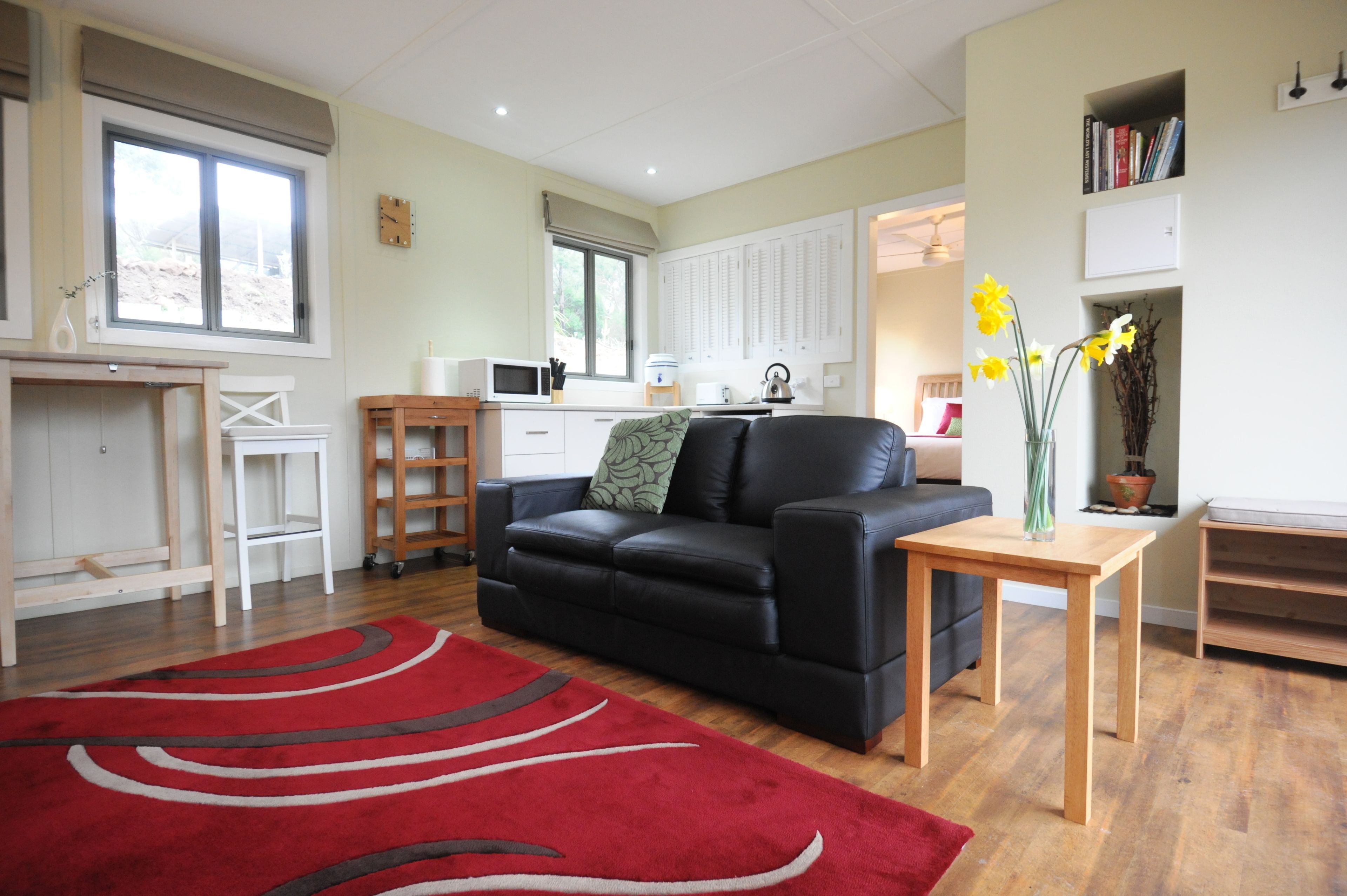 A modern living room with a black leather sofa, a red rug, a wooden coffee table, and a kitchen area in the background.