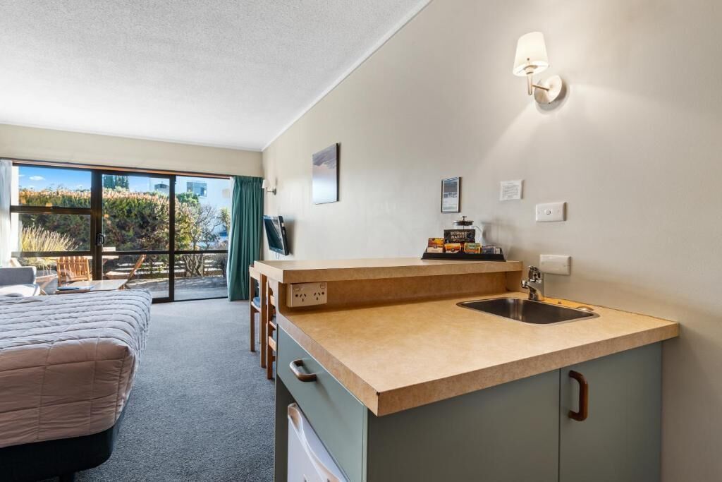 Quarto compacto com cama, pequeno espaço de cozinha com pia e uma grande porta de vidro deslizante que dá para uma área externa.