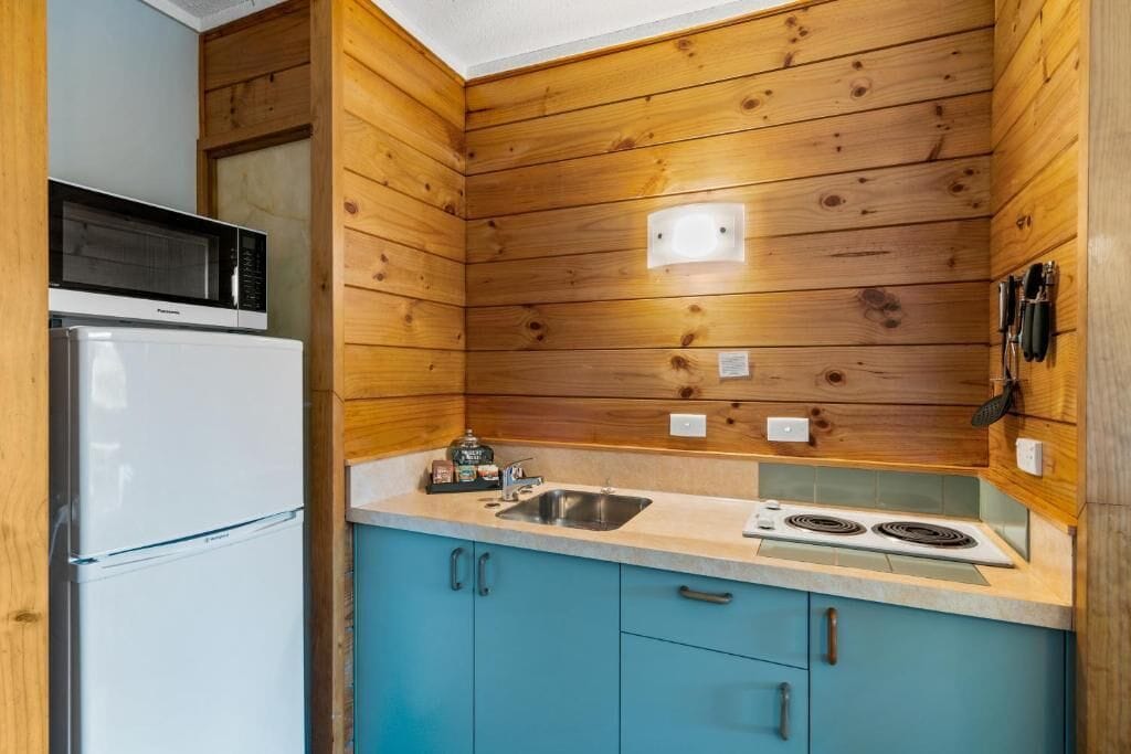 A compact kitchen with wooden walls, a white refrigerator, a microwave, a sink, and a stove.