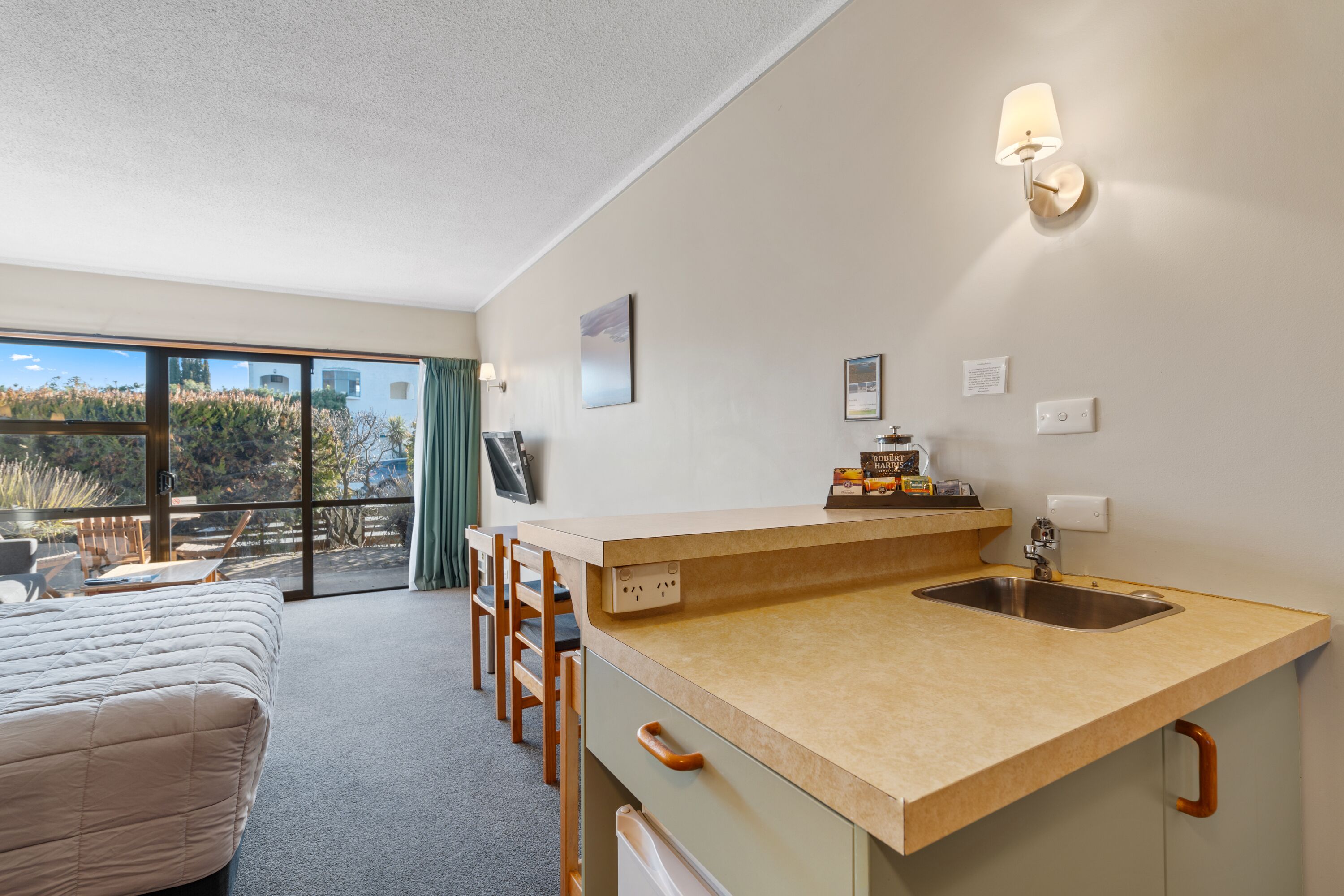 A compact hotel room with a kitchenette, a large window offering a view of the outdoors, and a bed with a striped cover.