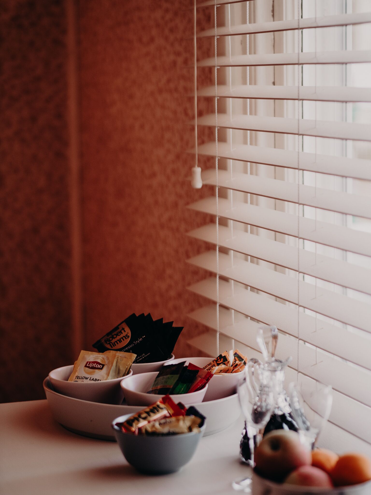 A table with bowls containing snacks and a bottle of wine, set against a window with blinds.