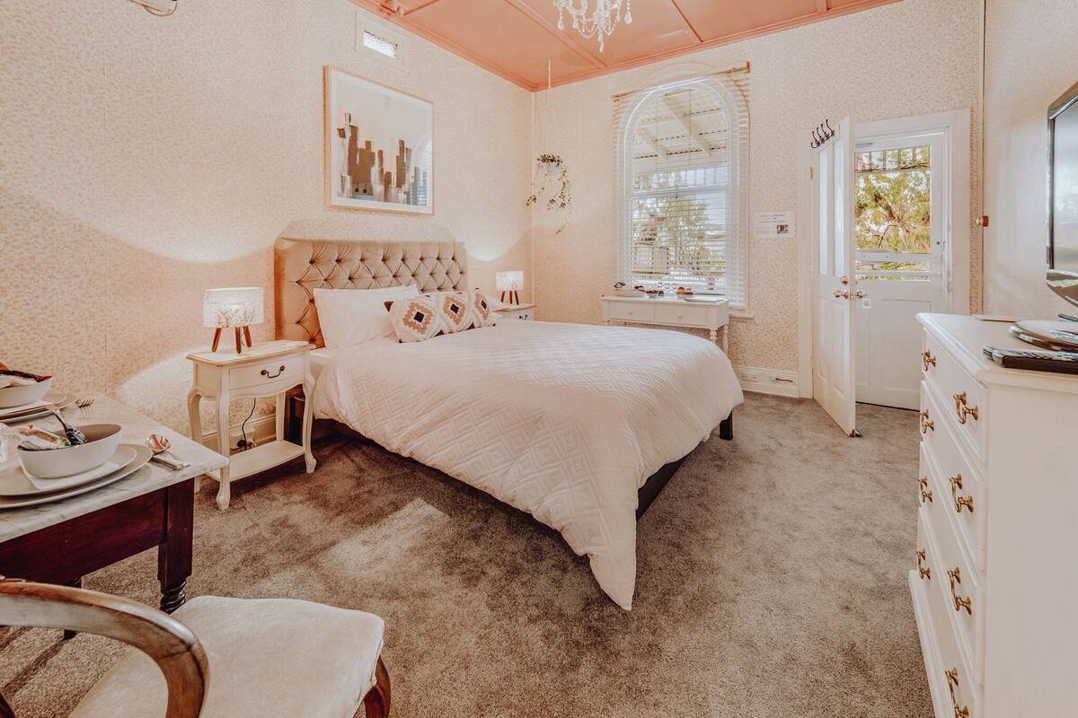 A bedroom with a large bed, a desk, a chair, a dresser, a chandelier, a window with blinds, and a framed picture on the wall.