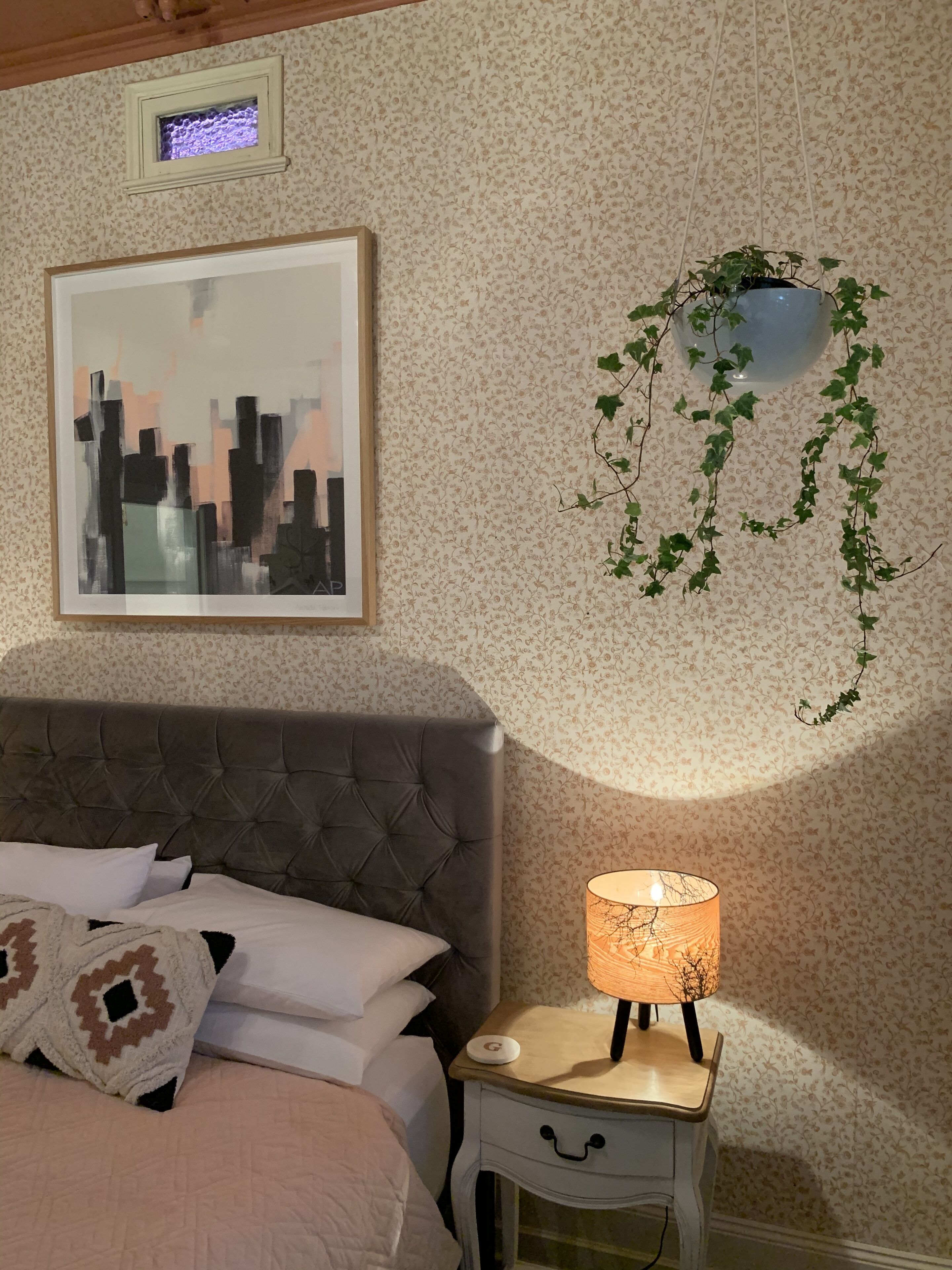A bedroom with a tufted headboard, a bed with white and patterned pillows, a bedside table with a lamp, a framed abstract cityscape painting, and a hanging plant.