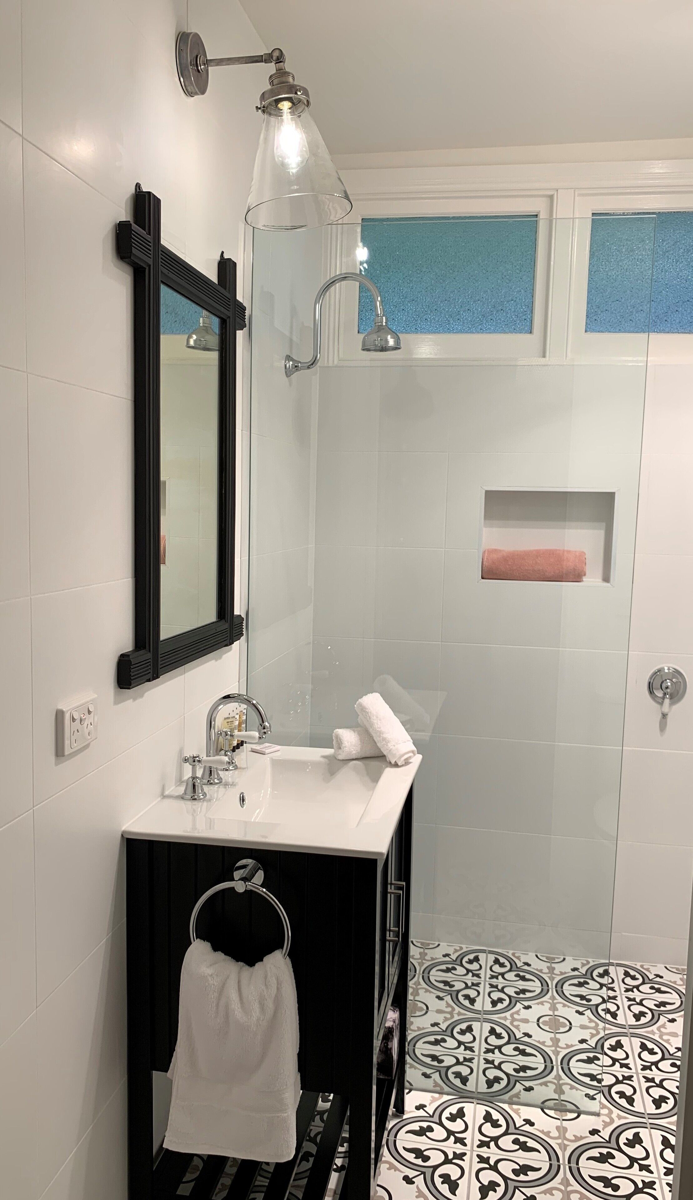 Een badkamer met een wit wastafelmeubel, een spiegel, een handdoekhouder, een douche met glazen deur en vloertegels met een patroon.