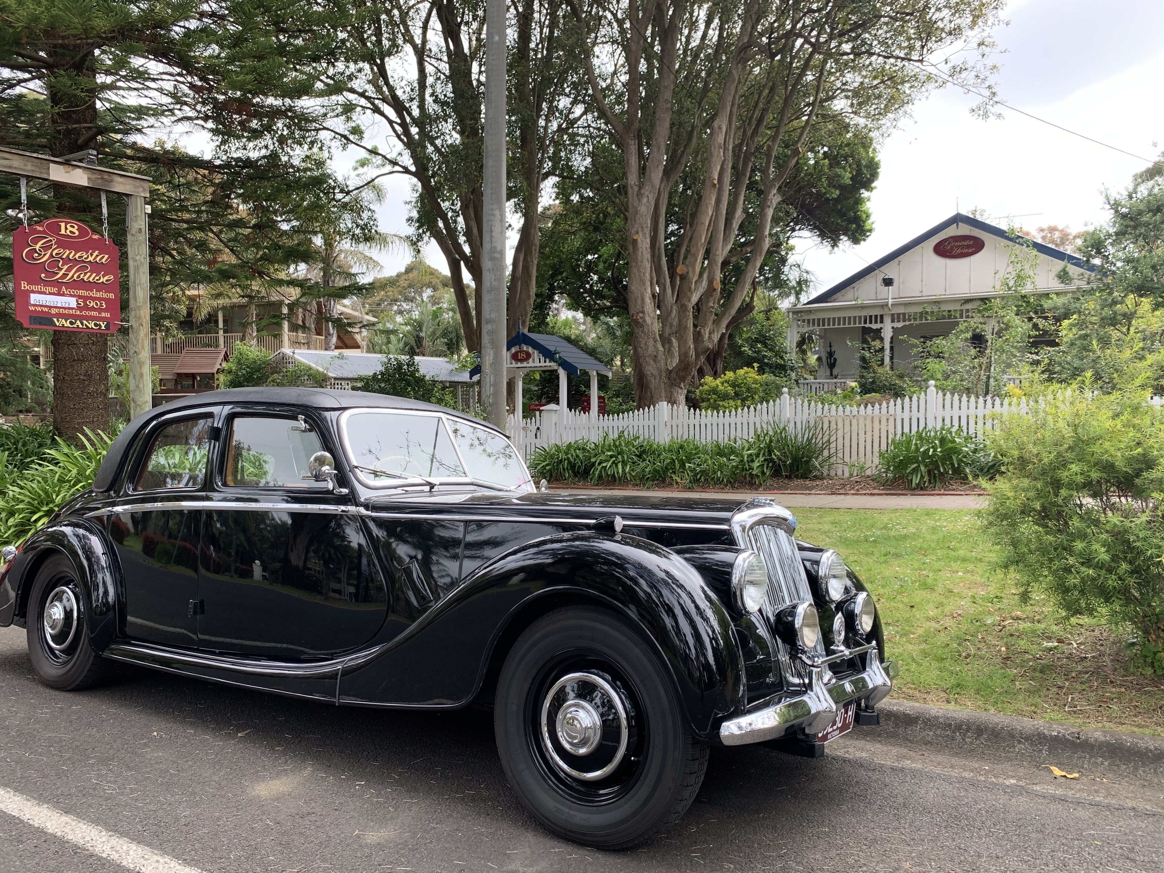 A vintage black car parked on a street with a sign for 'Genesta House Boutique Accommodation' and a 'VACANCY' sign.