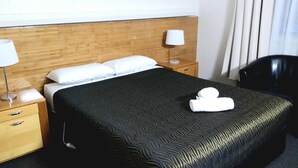A hotel room with a wooden headboard, a black patterned bedspread, white pillows, a bedside table with a lamp, and a black chair.