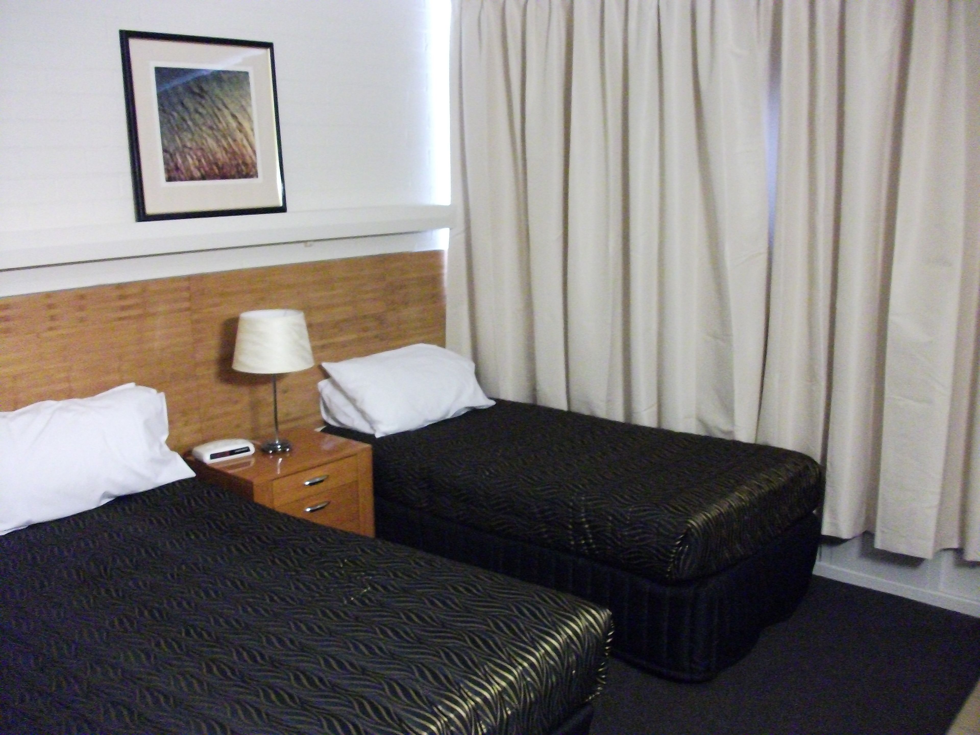 A hotel room with two beds, a nightstand with a lamp, a framed picture on the wall, and a window with curtains.