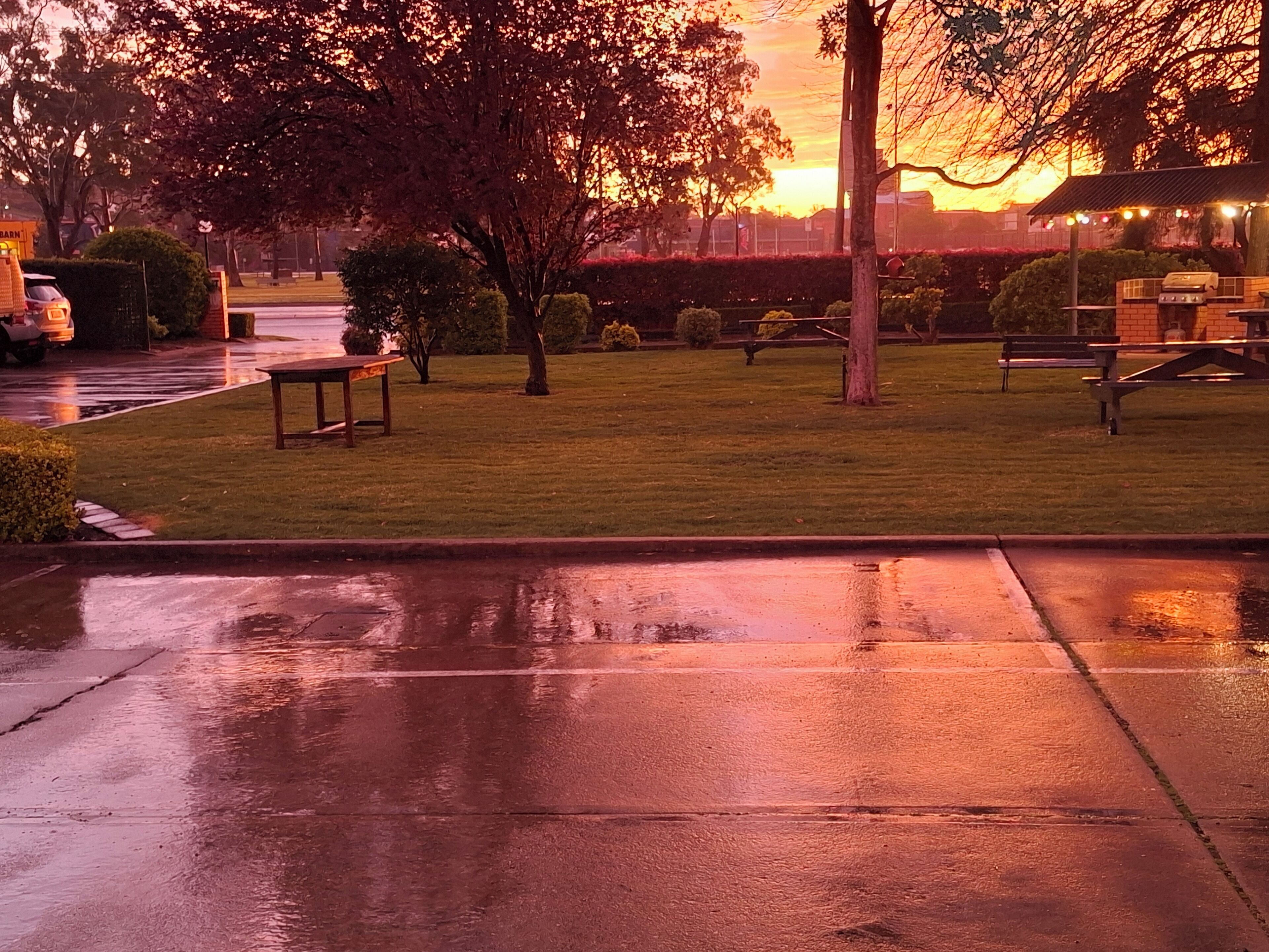A wet sidewalk with a bench and a picnic table in a park during sunset.