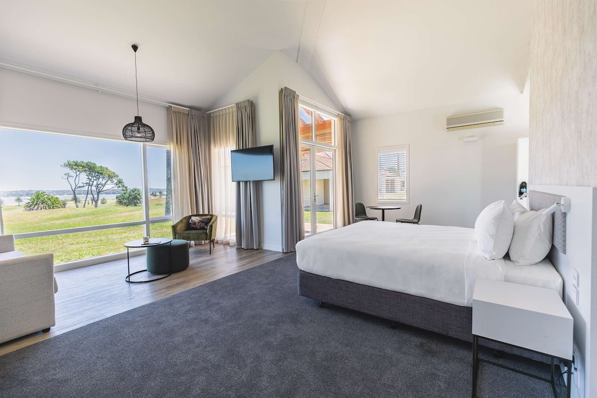 A modern hotel room with a large bed, a TV, a sofa, and a view of the outdoors.