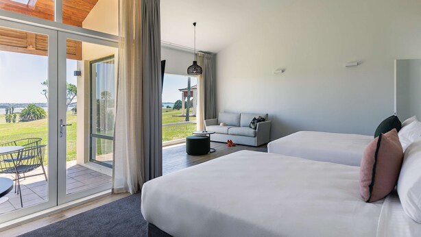 Habitación de hotel moderna con un amplio ventanal corredera que da a un espacio exterior, un sofá, dos camas y una lámpara colgante.