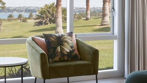 Un sillón verde con un cojín floral junto a una ventana que da a un área verde con palmeras y una masa de agua.