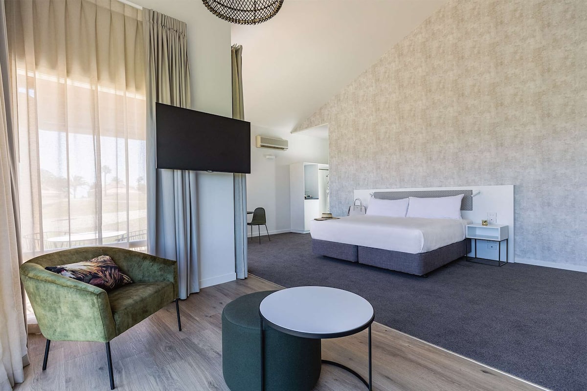 A modern hotel room with a large bed, a TV, a green armchair, and a small table.