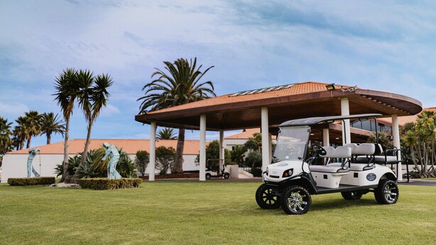 Un carrito de golf estacionado bajo un pabellón con palmeras y un edificio al fondo.