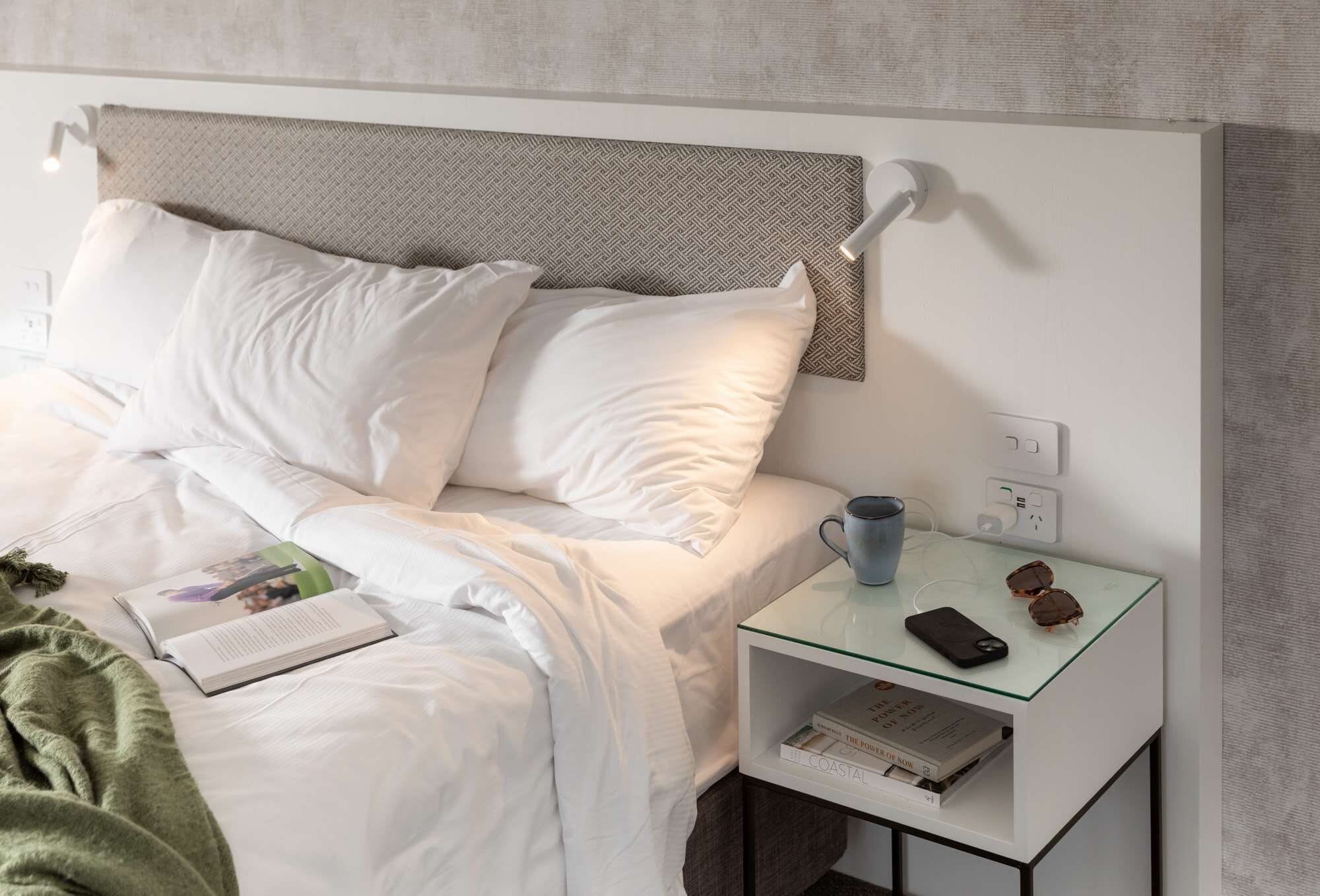 A neatly made bed with white linens, a bedside table with a glass top, a book, a mug, and a smartphone.