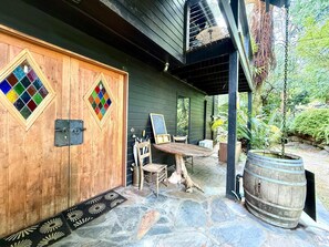 Un edificio de madera con dos puertas de vidrio emplomado, un patio de piedra, una mesa de madera y un barril grande.