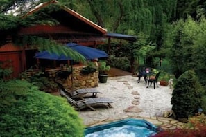 A rustic outdoor area with a stone patio, lounge chairs, a small pool, and a wooden structure with a thatched roof. - Forget Me Not Cottages (Warburton)