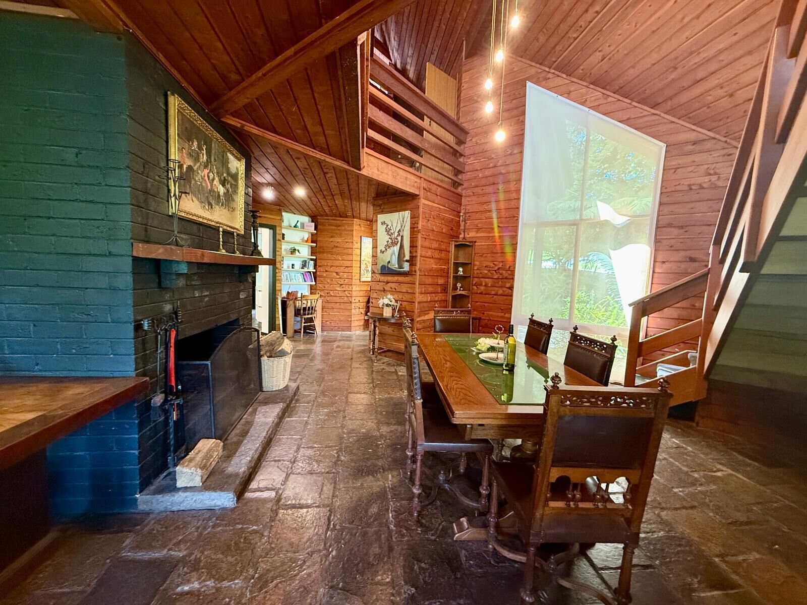 A cozy cabin interior with a fireplace, wooden walls, a dining table, and a large window.