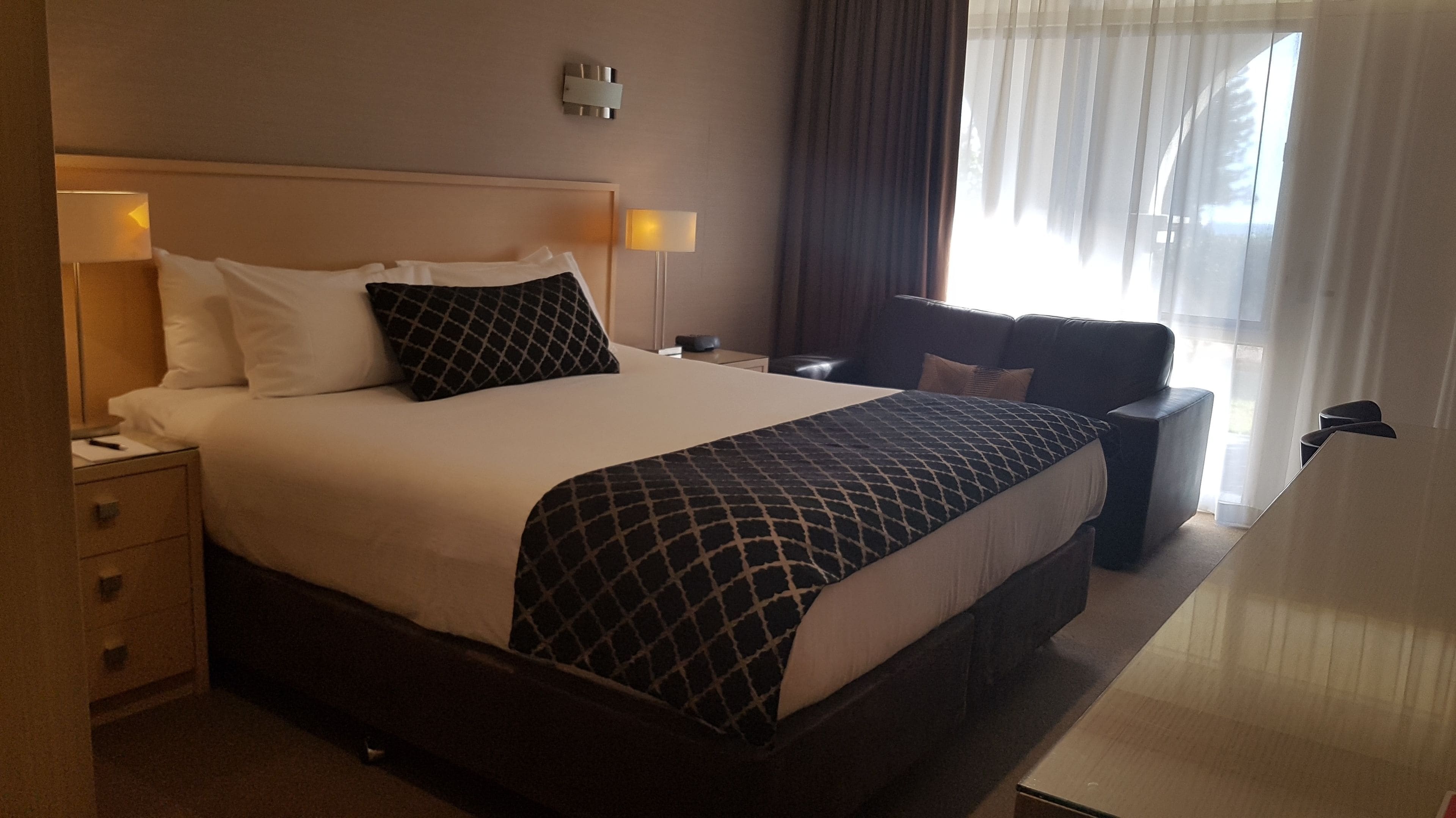 Une chambre d’hôtel avec un grand lit, deux tables de chevet, un bureau, une chaise et une fenêtre avec des rideaux.