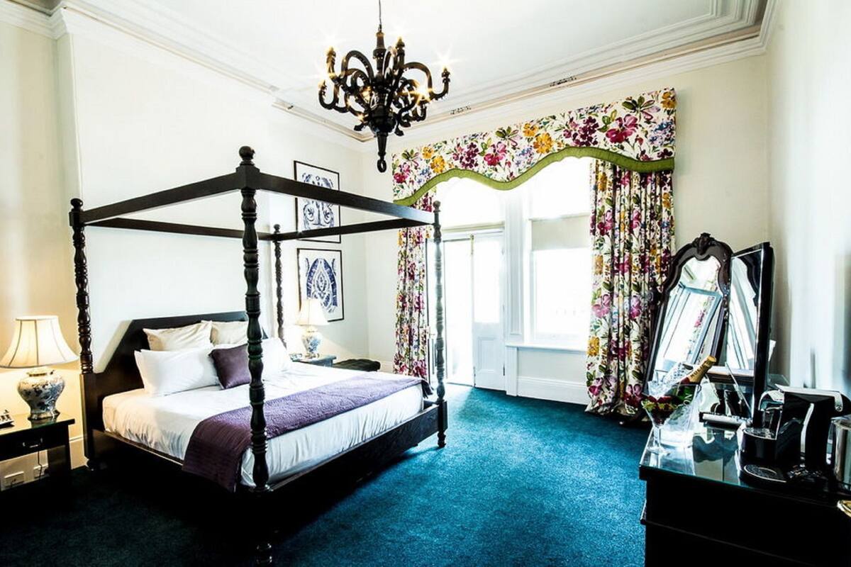 A four-poster bed with a dark finish, a chandelier, a floral curtain, and a mirror.
