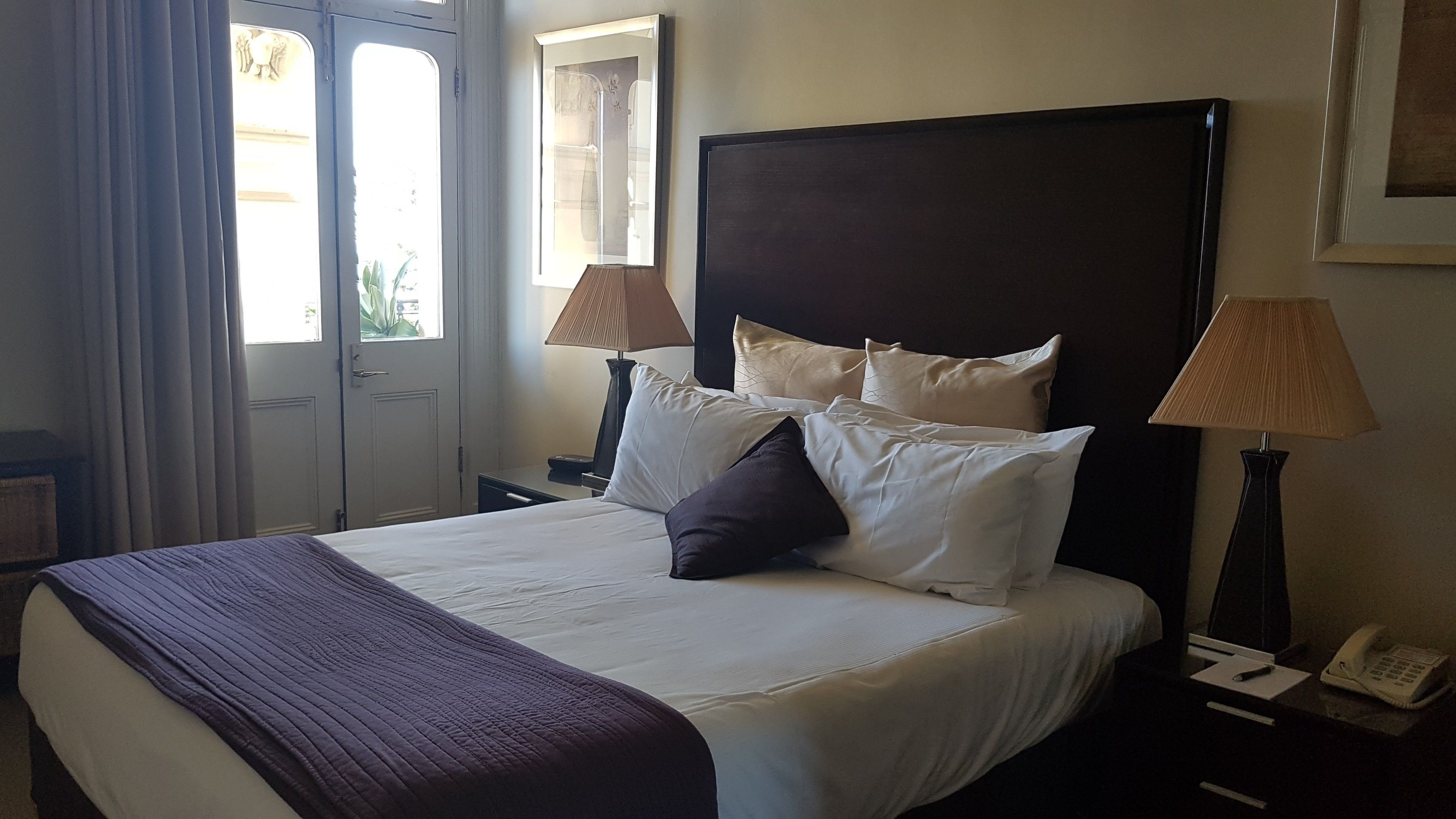 Quarto de hotel com uma cama grande, duas luminárias de cabeceira, mesa de cabeceira com telefone e uma janela com cortinas.