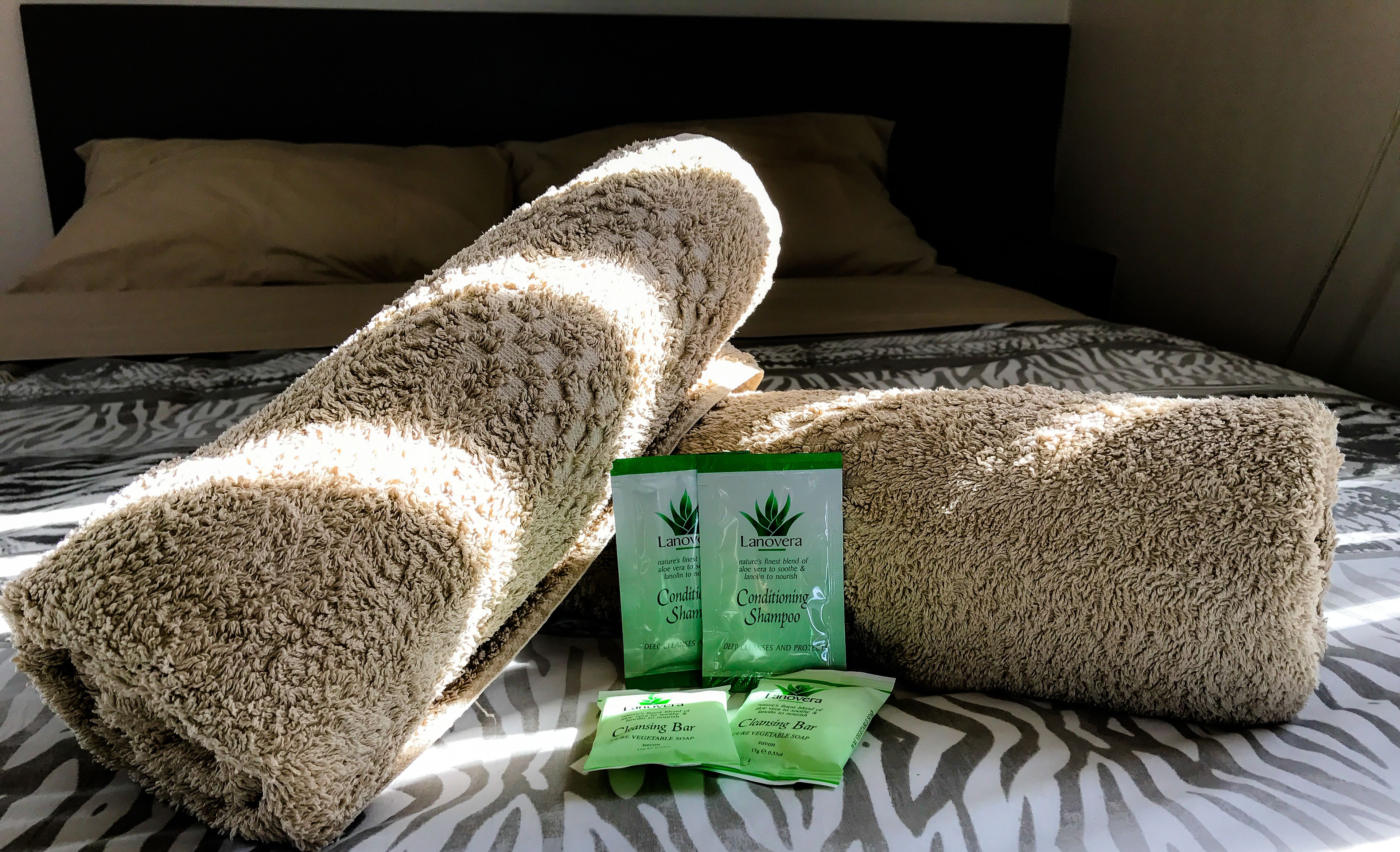 A bed with a zebra-patterned bedspread, two rolled-up towels, and two packets of Lanavera conditioning shampoo and cleansing bar.