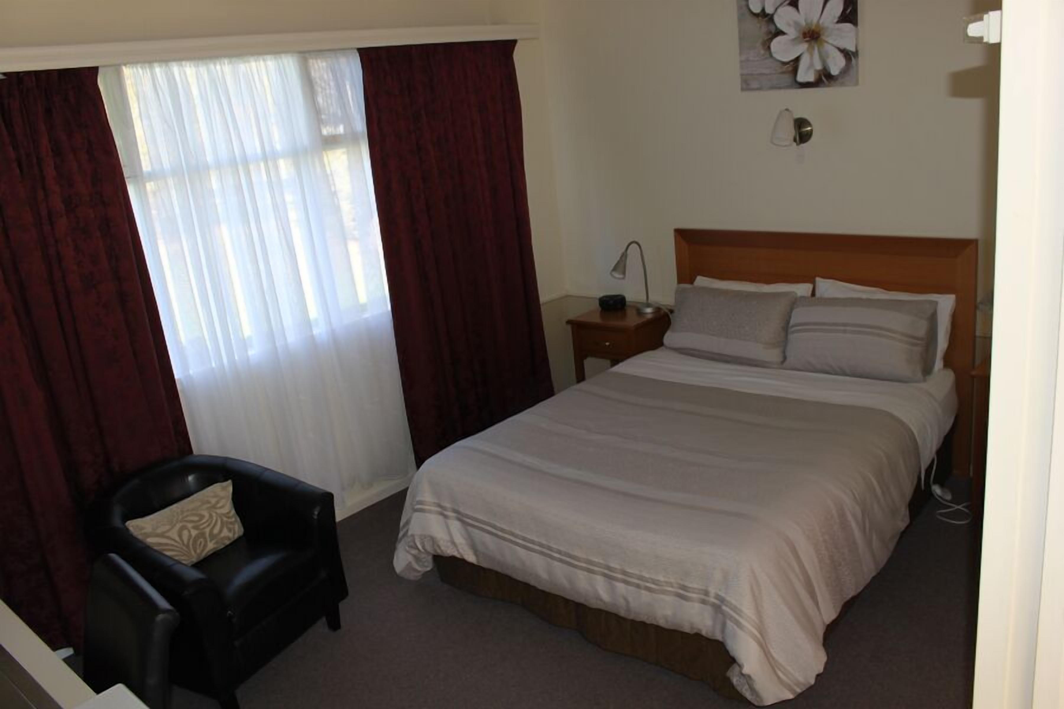 A hotel room with a large bed, a chair, a nightstand, a lamp, and a wall with a flower painting.