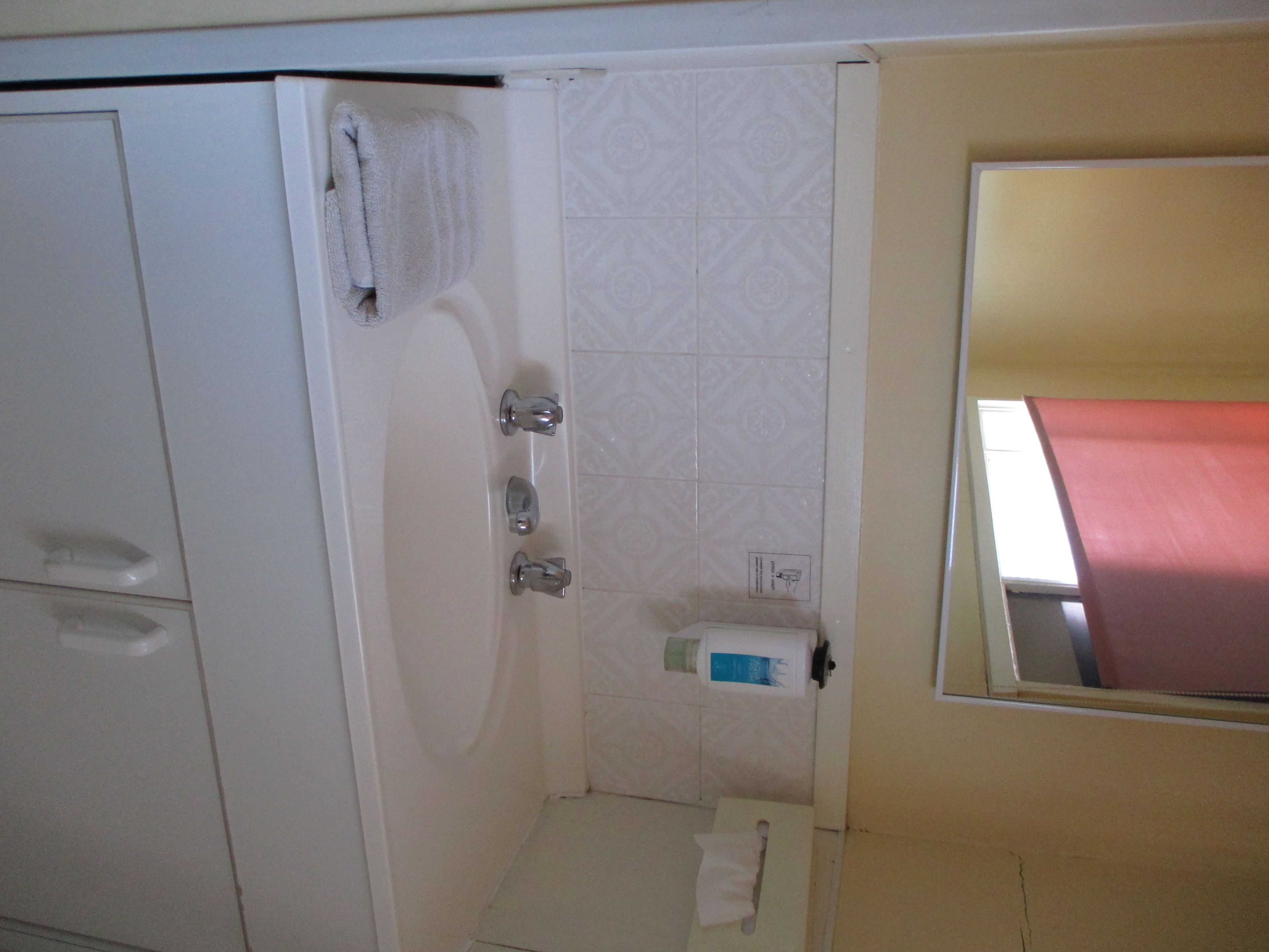 Un pequeño espacio de ducha cerrado con un muro de azulejos blancos, una toalla colgada en la pared y un inodoro con dispensador de toallas de papel.