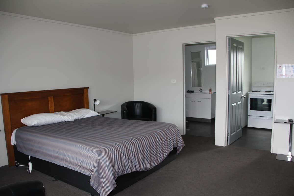 A bedroom with a bed, a chair, a desk, and an open door to a kitchenette.