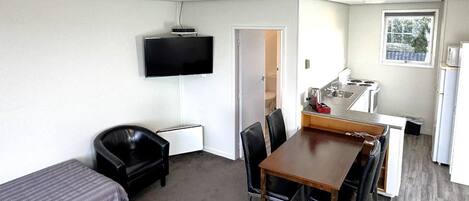 A compact room with a single bed, a desk with a chair, a television mounted on the wall, and a kitchenette area.