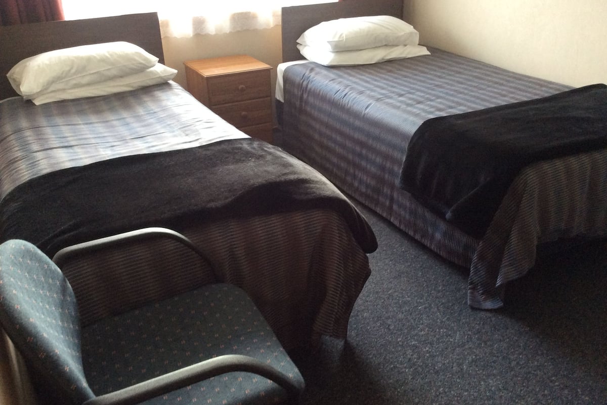 A hotel room with two single beds, a small wooden nightstand, a chair, and a window with curtains.