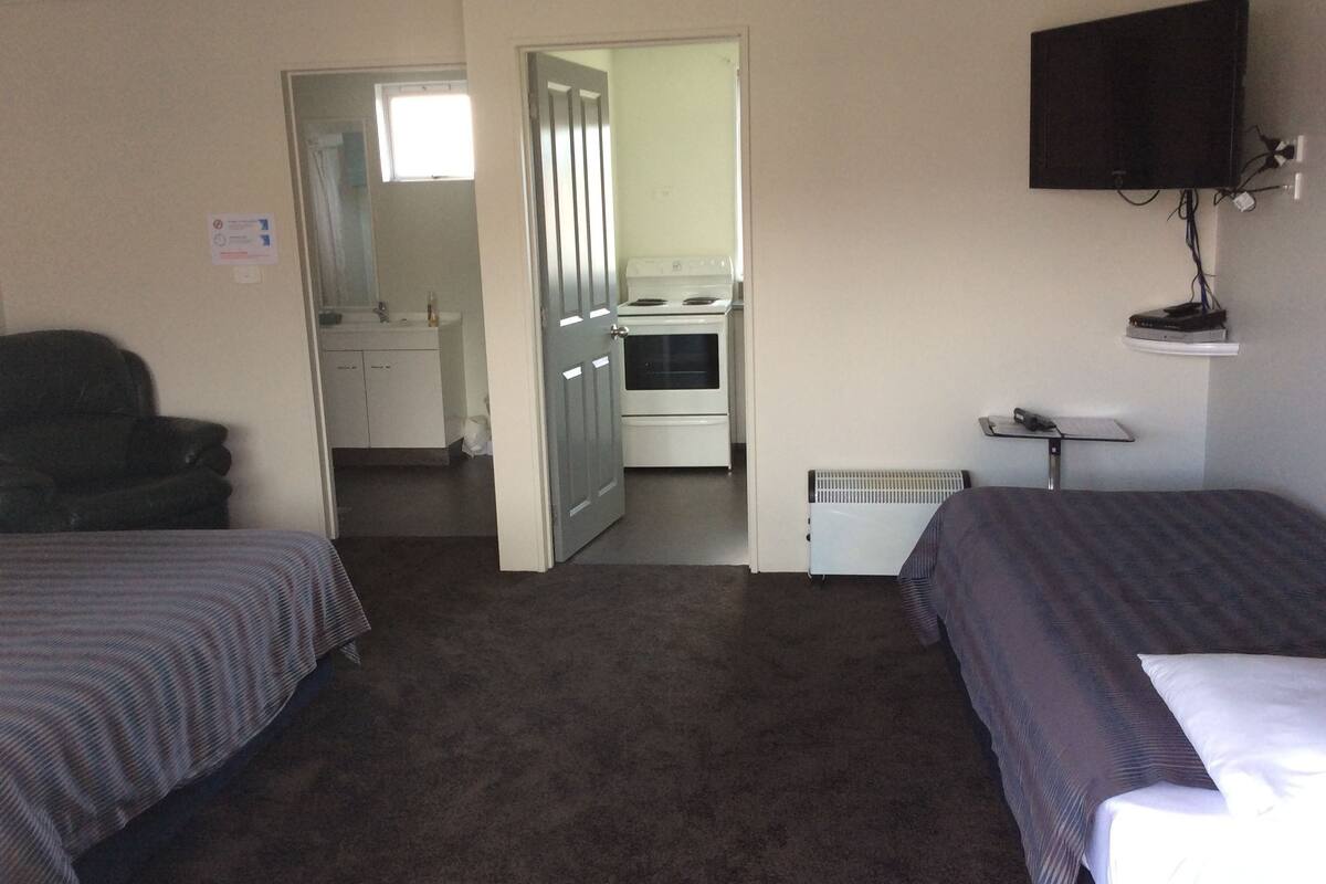 A hotel room with two beds, a TV, a desk, a chair, a bathroom with a sink and mirror, and a kitchen with a stove and oven.