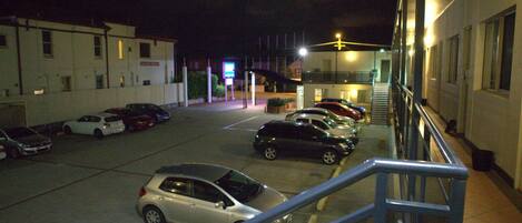 Un estacionamiento con varios coches aparcados, un edificio con una escalera y un cartel al fondo.