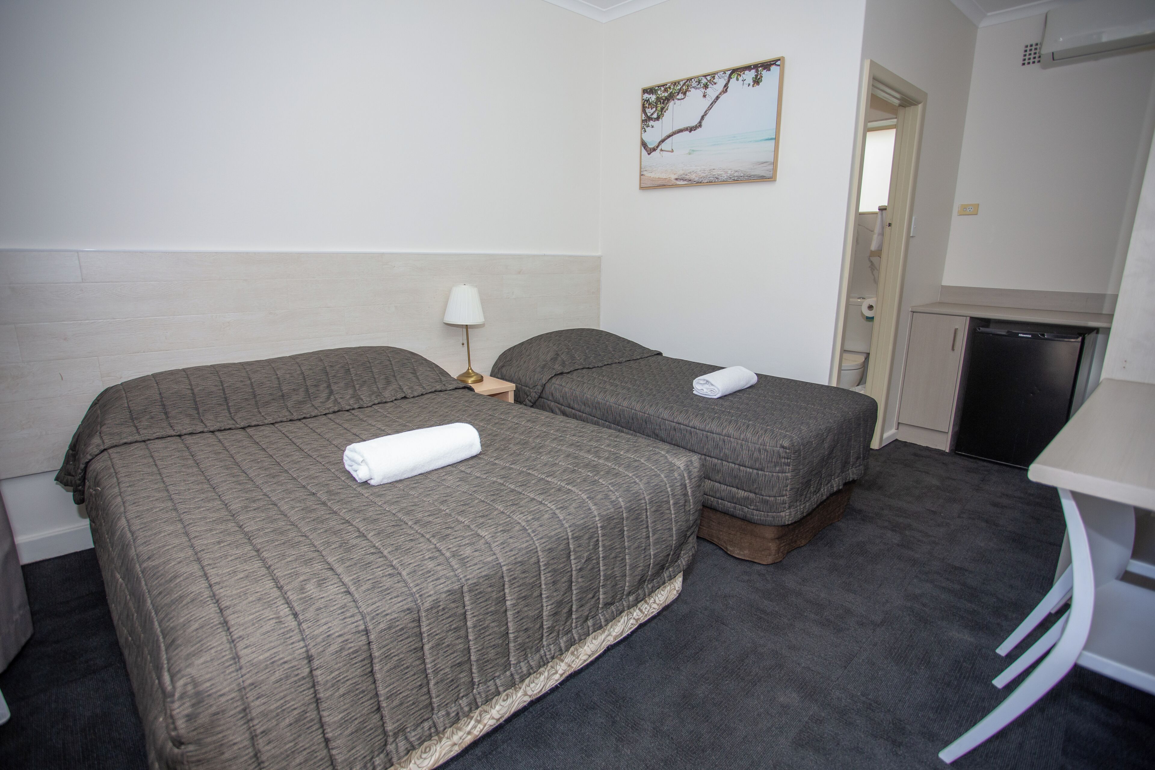 A hotel room with two beds, a small table, a lamp, a painting on the wall, and a kitchenette.