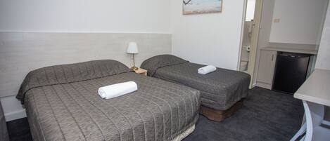 A hotel room with two beds, a small table, a lamp, a painting on the wall, and a kitchenette.