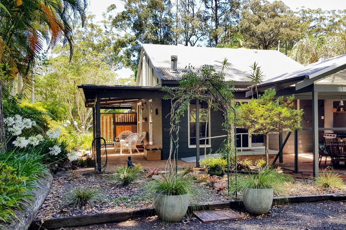 A modern house with a covered patio, a garden with various plants, and a large tree in the background.