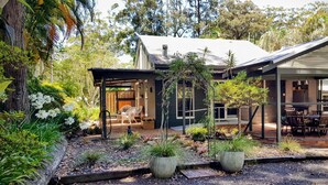 Una casa moderna con un porche cubierto, un jardín con diversas plantas y un árbol grande al fondo.