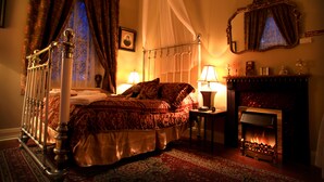 Ein Schlafzimmer mit einem Himmelbett, einem Nachttisch mit Lampe, einem Kamin und einem Spiegel an der Wand.