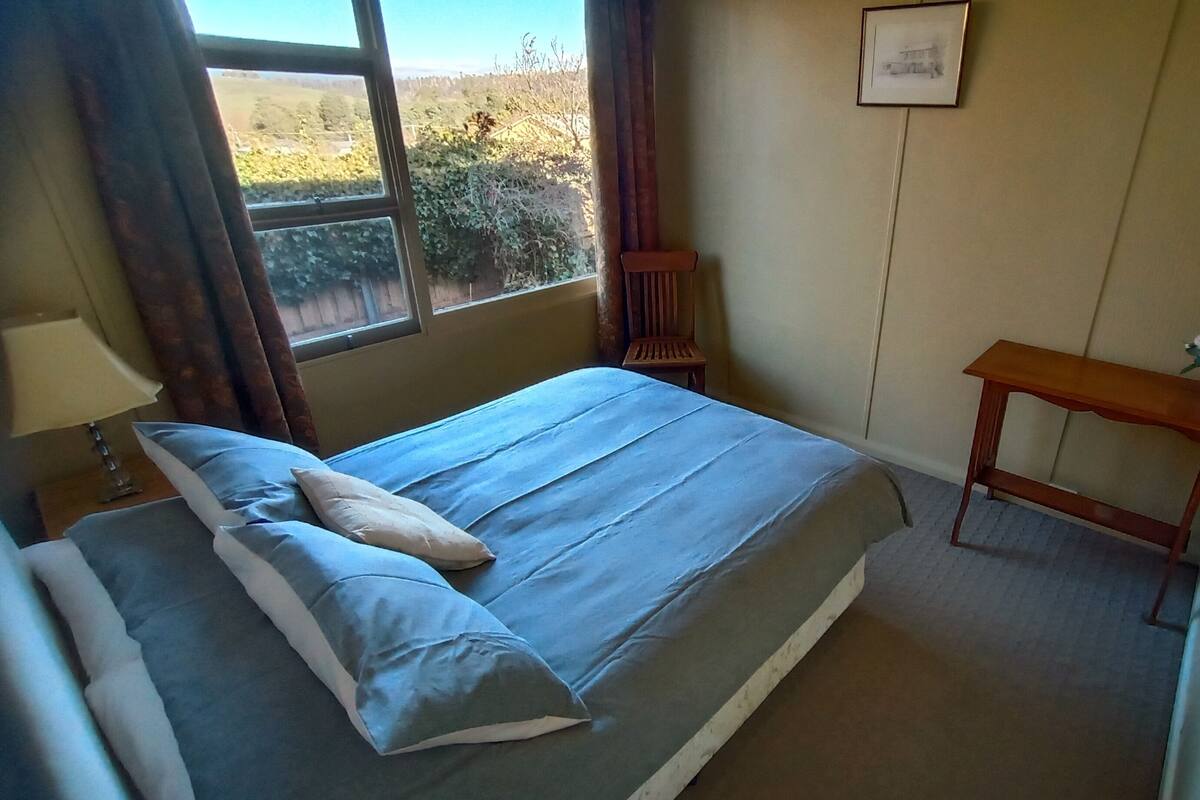 Ein Schlafzimmer mit Bett, Nachttisch, Fenster mit Vorhängen, Stuhl, kleinem Tisch mit Vase und einem gerahmten Bild an der Wand.
